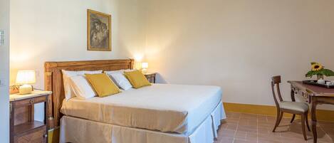 Una cama bien hecha con sábanas blancas y almohadones amarillos, una mesita de noche de madera con una lámpara, un cuadro en la pared y una pequeña mesa con un jarrón de girasoles.