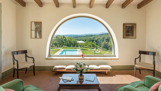 Ein Zimmer mit einem großen Bogenfenster mit Blick auf ein Schwimmbecken und einen Garten. Zwei Stühle und ein Tisch mit einem Blumenstrauß sind vorhanden.