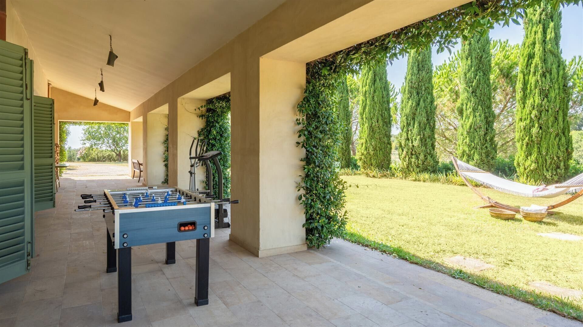 An outdoor patio with a foosball table, a hammock, and a view of a grassy area with tall trees.