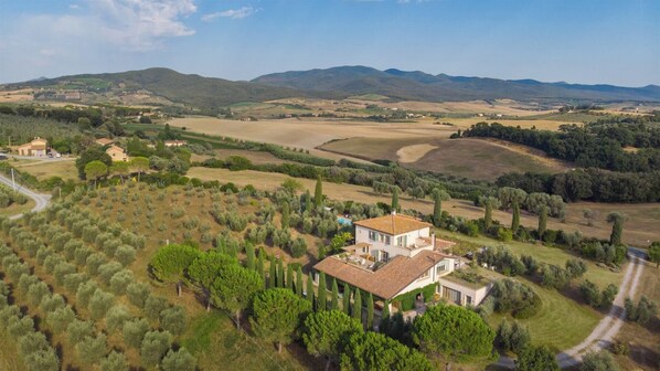 A large house with a tiled roof surrounded by olive groves and vineyards, set in a hilly landscape.