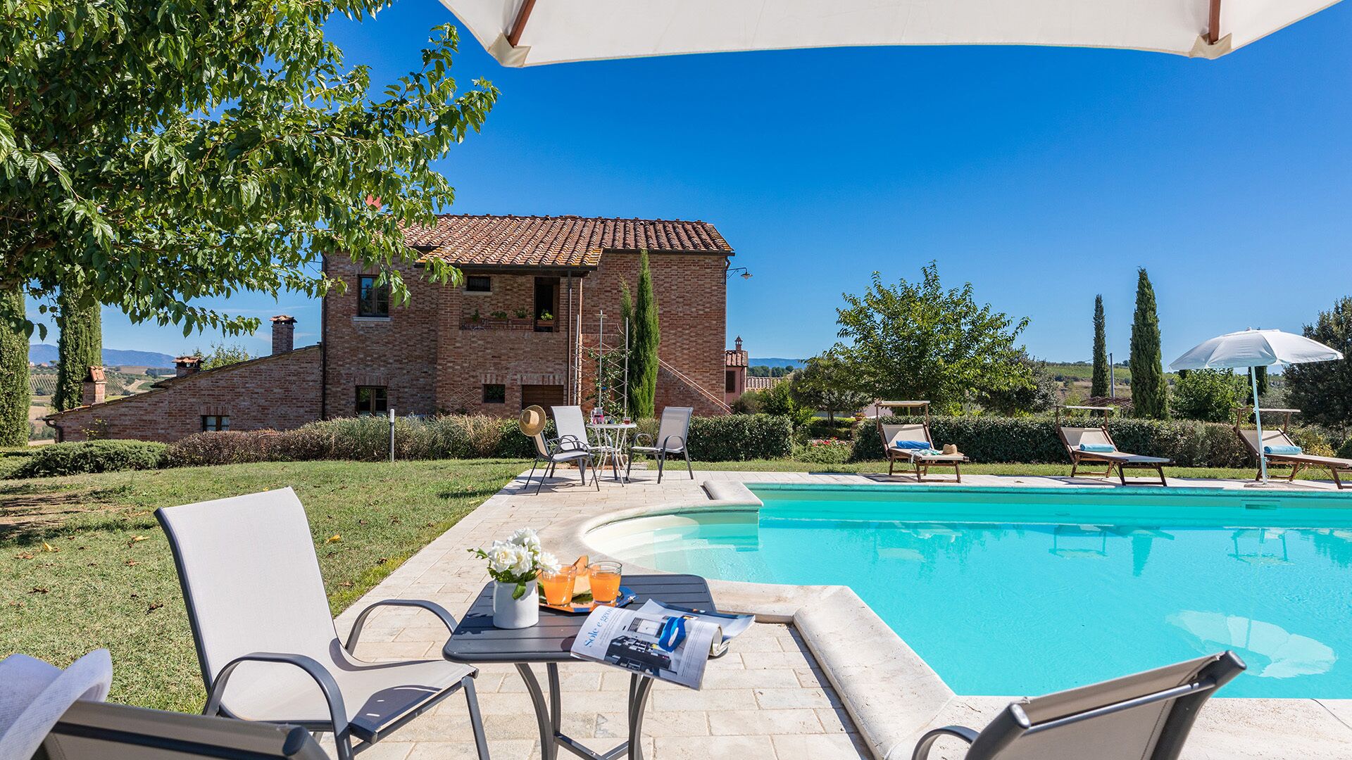 Un espace aménagé au bord de la piscine, avec des chaises longues, une table où sont servis des boissons et une vue sur un bâtiment en briques.