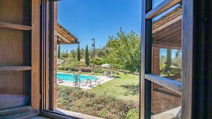 Vista d'una piscina amb gandules i una zona enjardinada, visible a través d'una finestra de fusta.