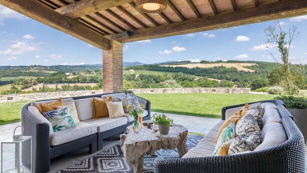 An outdoor seating area with wicker furniture, a stone table, and a view of rolling hills. - Villa Le Fontane 12, Emma Villas (Todi)