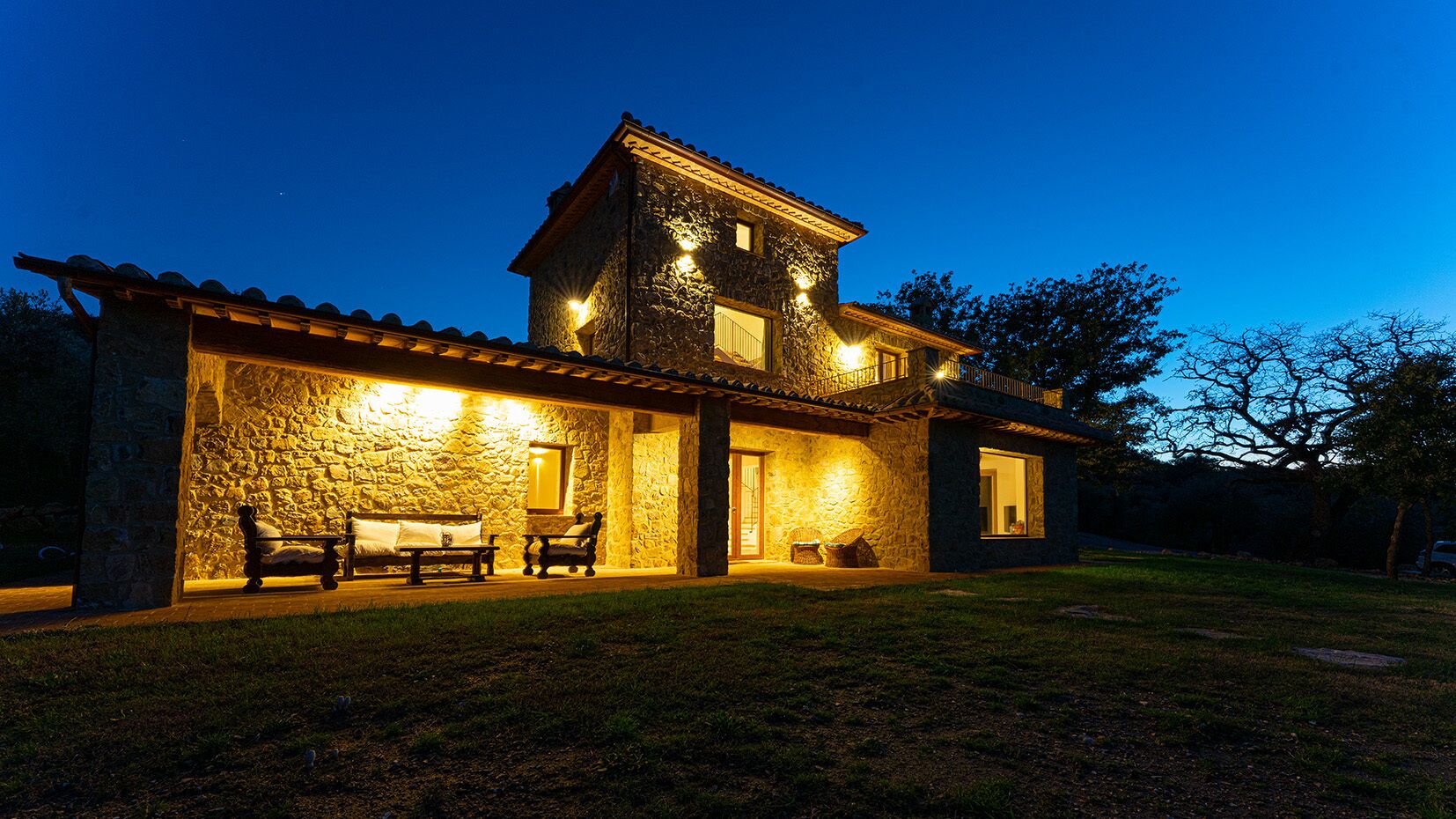 Una casa de piedra con balcón, un porche con banca y mesa, y una entrada bien iluminada.