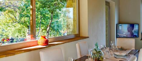 A dining area with a table set for two, a window with a view of trees, and a television on the wall.
