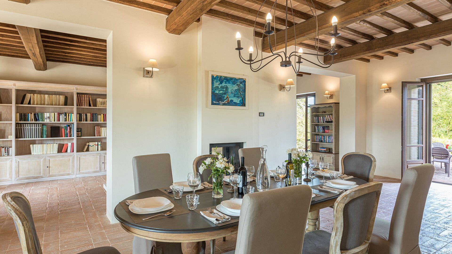 A dining area with a round table set for four, surrounded by chairs. There are bookshelves on either side of the room, and a chandelier hangs from the ceiling.