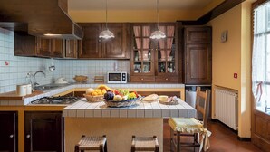 Een keuken met houten kastjes, een betegelde spatwand, een centraal eiland met fruit en een eethoek met stoelen.
