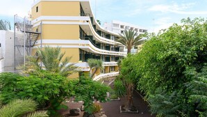 Un edificio moderno con fachada curva, varios balcones y un jardín ajardinado con plantas diversas y una palmera.