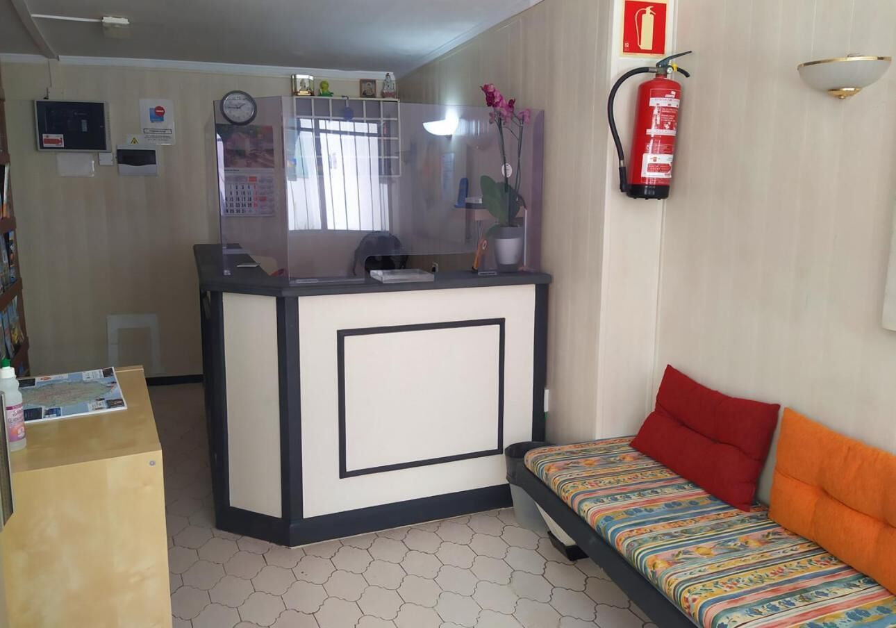 A reception area with a desk, a bed with colorful bedding, a fire extinguisher, and a clock on the wall.