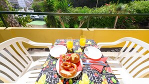 Una mesa en el balcón dispuesta con alimentos para el desayuno, dos vasos de jugo de naranja y vista a un paisaje urbano con áreas verdes.