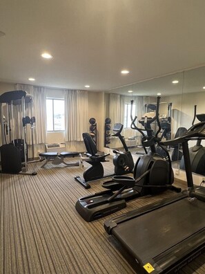 Academia de hotel com equipamentos de cardio, máquinas de peso e um espelho grande.
