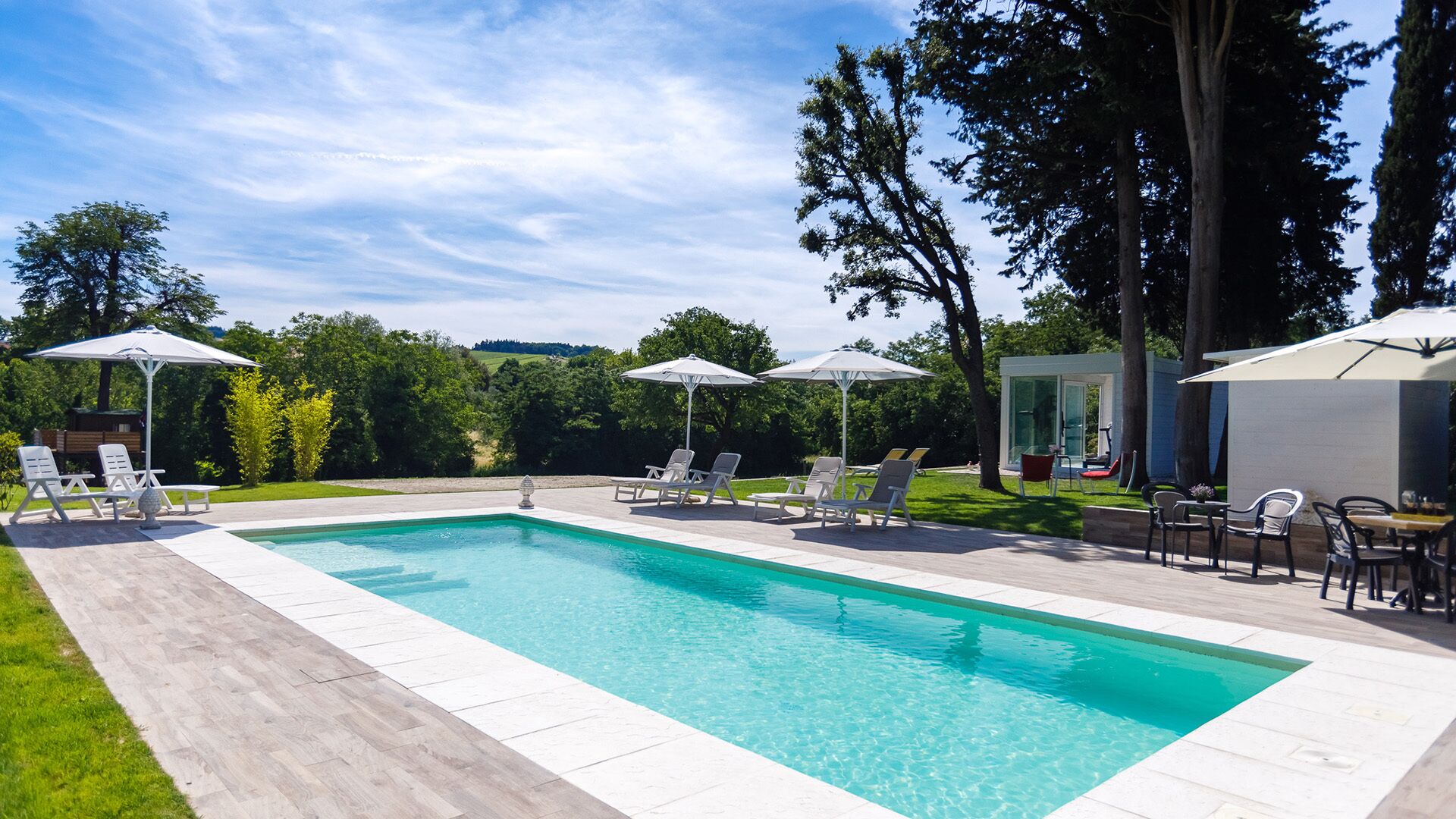 Une piscine rectangulaire à l’eau d’un bleu limpide, entourée d’une terrasse carrelée. Des chaises longues et des parasols sont disposés autour de la piscine, et un petit bâtiment aux portes vitrées se trouve à l’arrière-plan.