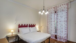 A bedroom with a bed, bedside lamps, a radiator, and a window with patterned curtains.