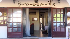Entrada de hotel con puertas dobles, un letrero que dice 'Bravaville' y un interior visible con una mesa y sillas.