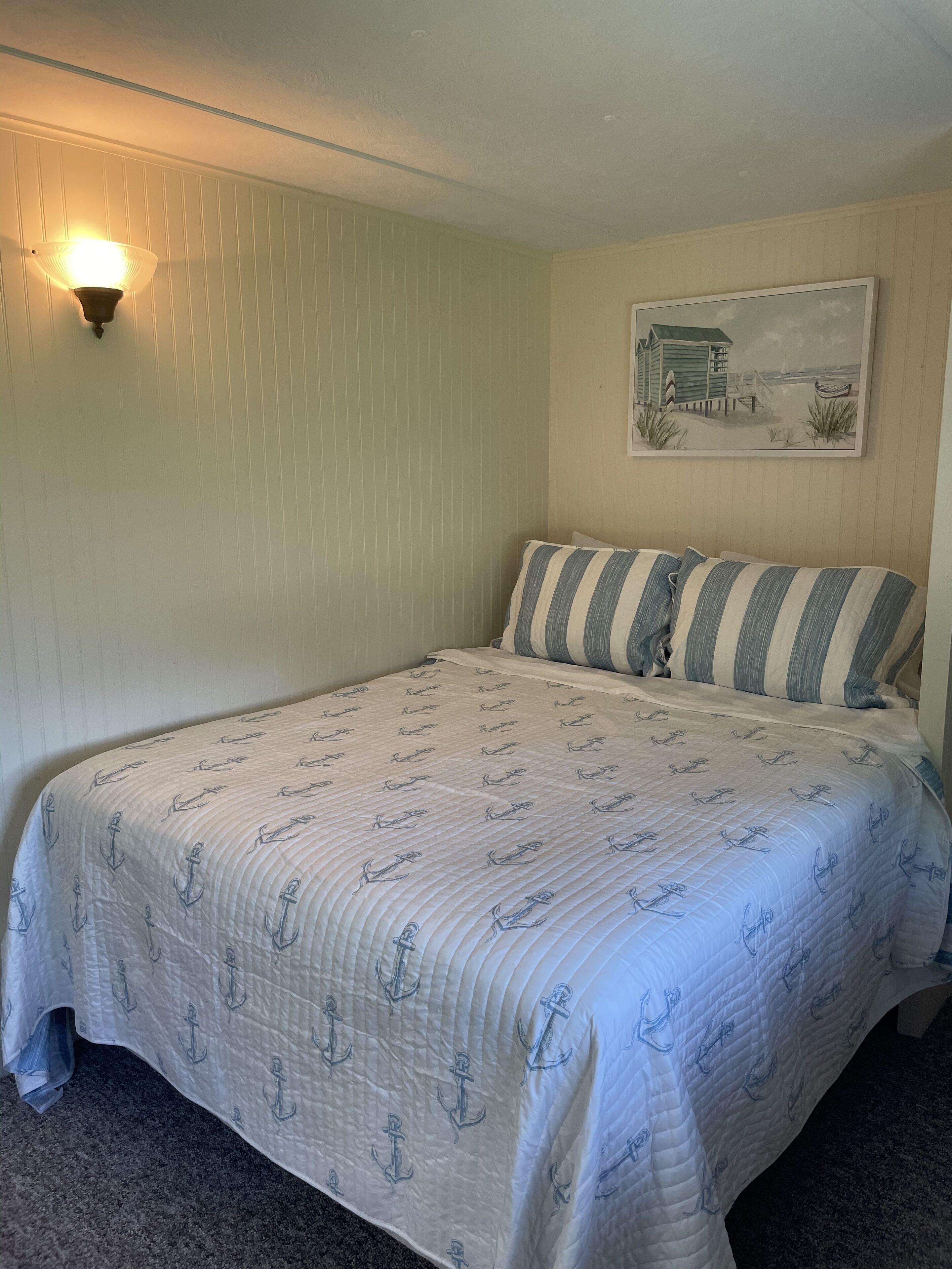 A bedroom with a bed, striped pillows, a wall-mounted lamp, and a beach-themed painting.