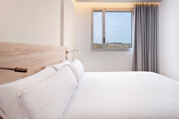 A neatly made bed with white linens and a wooden headboard in a hotel room with a large window and curtains. - B&B Hotel Murcia (Murcia)