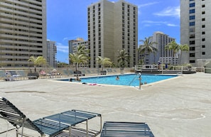 Un área de piscina en la azotea con sillones reclinables, una piscina y edificios altos al fondo.