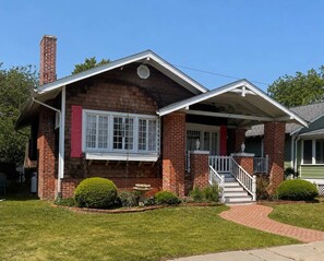 A two-story brick house with a front porch, a chimney, and a small garden. - Hummingbird Cottage with Park Views, Pet-Friendly (Cape Charles)