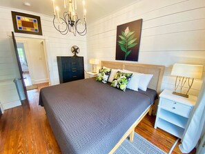 A bedroom with a large bed, a bedside table with a lamp, a dresser, a fan, and a painting on the wall. - Hummingbird Cottage with Park Views, Pet-Friendly (Cape Charles)