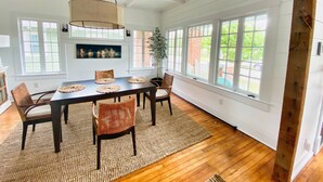 A dining room with a large table, four chairs, a hanging light fixture, and large windows with a view of greenery outside. - Hummingbird Cottage with Park Views, Pet-Friendly (Cape Charles)