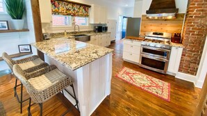 A spacious kitchen with a large island, wooden flooring, and a brick accent wall. - Hummingbird Cottage with Park Views, Pet-Friendly (Cape Charles)