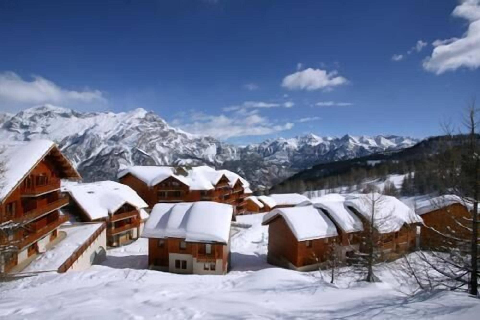 A cluster of wooden cabins with snow-covered roofs, set against a backdrop of snow-capped mountains and a clear blue sky.