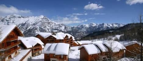 A cluster of wooden cabins with snow-covered roofs, set against a backdrop of snow-capped mountains and a clear blue sky.
