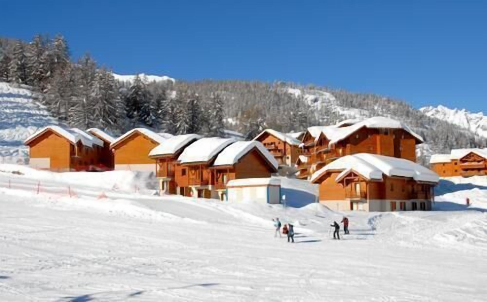 Una hilera de chalets de madera con techos nevados, frente a un fondo de árboles y montañas cubiertas de nieve.
