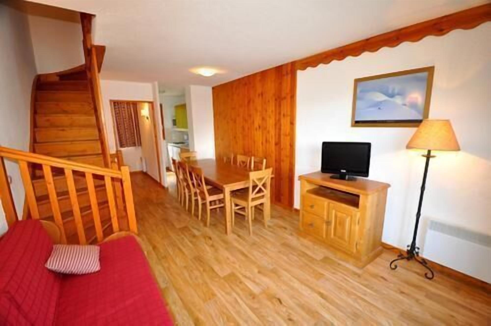 A wooden-floored living area with a red sofa, a dining table with chairs, a TV on a wooden cabinet, and a staircase.