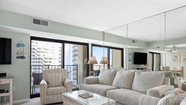 Una sala de estar con sofá, sillón y una mesa de centro, una televisión, y un balcón con vistas a edificios.