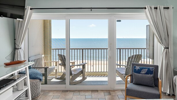 Un balcón con vista a la playa, dos mecedoras, una mesa y un televisor a la izquierda.
