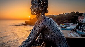 En skulptur af en menneskelig figur med et mønstret design, der kigger ud over en strand med solnedgang i baggrunden.