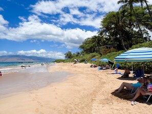 Uma praia com pessoas relaxando sob guarda-sóis, céu claro e vista para o oceano.