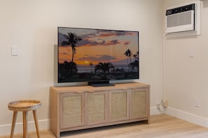 Uma televisão de tela plana em um armário de madeira com painel frontal trançado, um banquinho de vime e um aparelho de ar condicionado na parede.