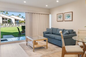 Un salon moderne avec une table basse en verre, un canapé gris et une vue sur une cour aménagée avec du mobilier d’extérieur.
