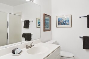 A bathroom with a white sink, mirror, and toilet. There are towels hanging on the wall and a framed picture above the toilet.