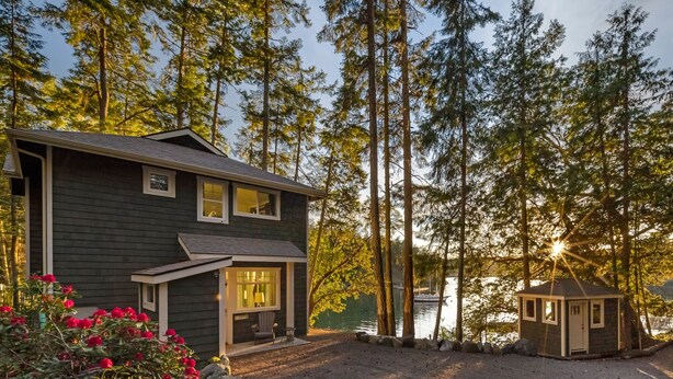 Une maison avec une allée en gravier, un petit abri de jardin et une jetée qui s’avance dans un lac bordé de grands arbres.