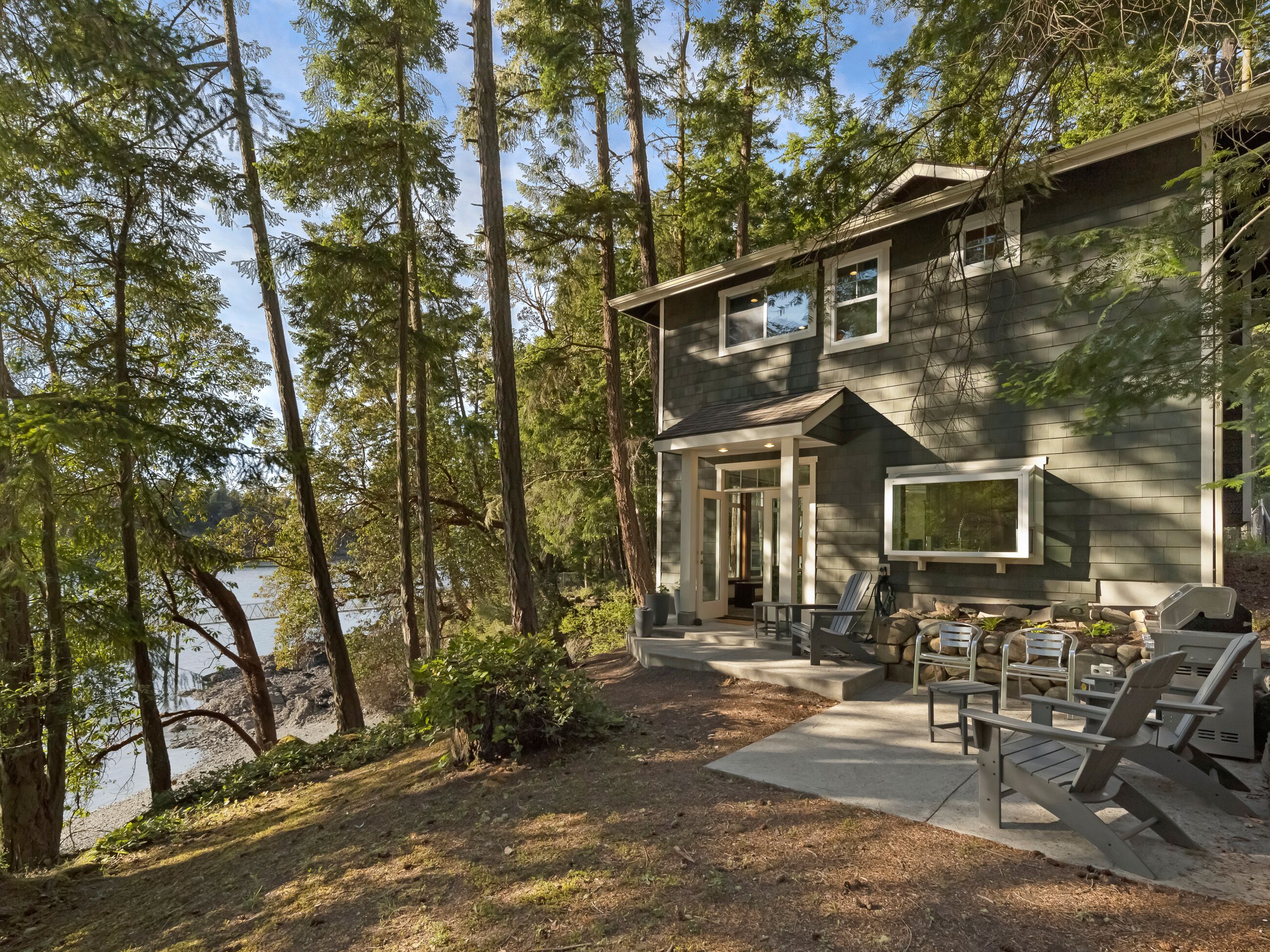 Une maison moderne dotée d’une terrasse, d’un espace salon extérieur et offrant une vue sur un lac entouré de grands arbres.