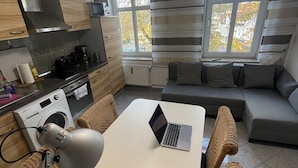 Kompakti keittiö, jossa on mikroaaltouuni, vedenkeitin ja leivänpaahdin. Kannettava tietokone on pöydällä, ja olohuoneessa on sohva, josta on näkymä ulkona oleviin puihin.