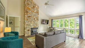Un vaste salon doté d’une cheminée en pierre, d’un canapé gris, de fauteuils turquoise et d’une grande fenêtre offrant une vue sur la verdure.