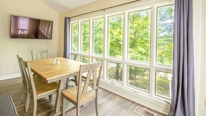 Un coin repas avec une table et des chaises en bois, de grandes fenêtres avec des rideaux et un téléviseur à écran plat fixé au mur.