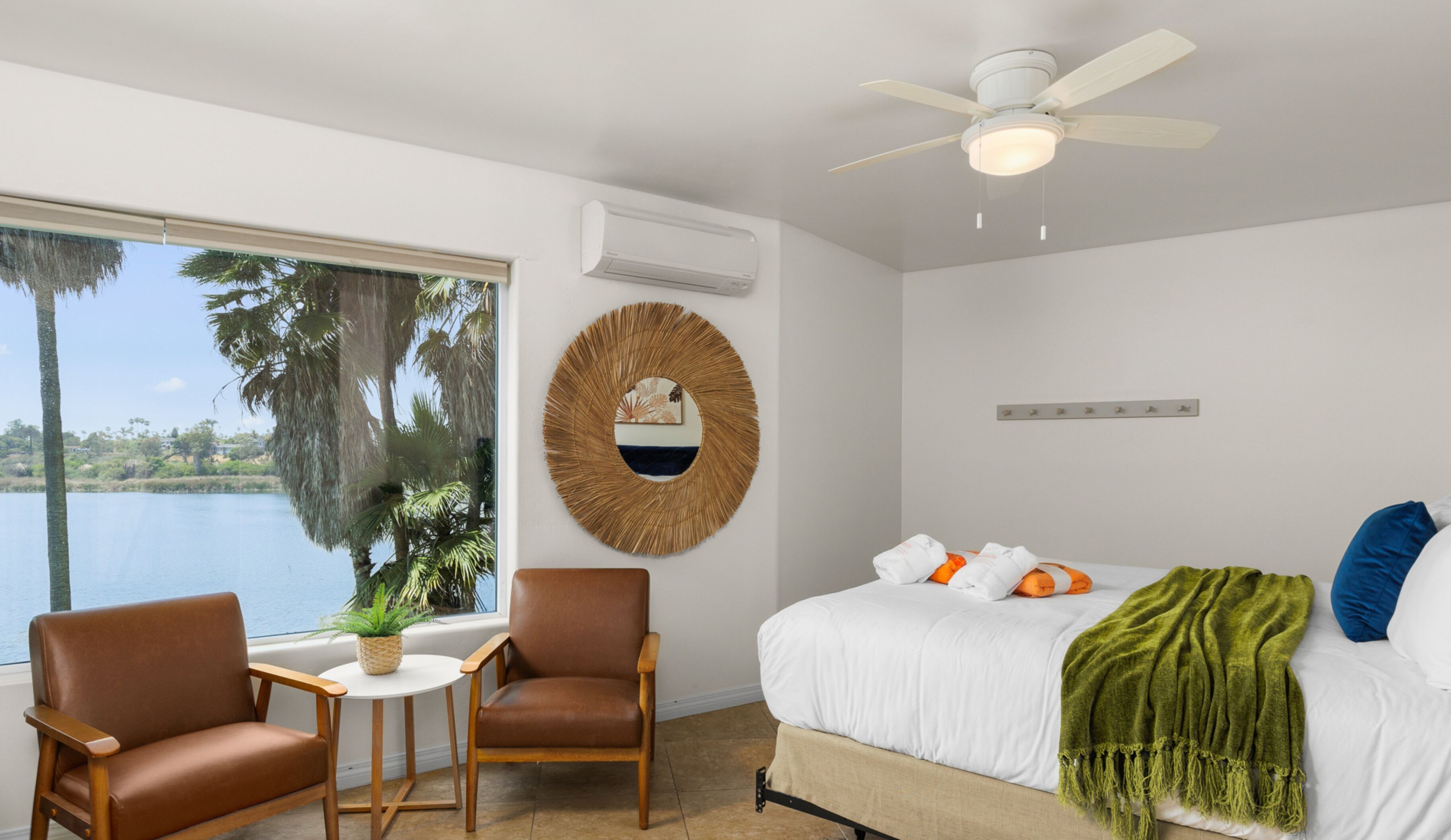 Una camera da letto con un letto, una ventola a soffitto, un decoro circolare a parete, una finestra vista palme e acqua, e due sedie.