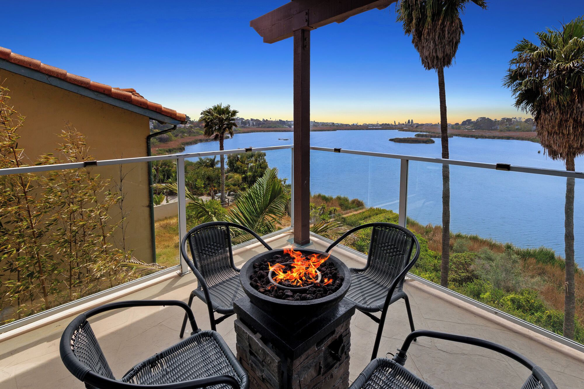 A balcony with a fire pit, two chairs, and a view of a lake and distant buildings.