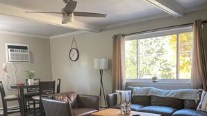 A living room with a sofa, a coffee table, a dining table with chairs, a ceiling fan, and a window with curtains. - Adorable 2 Bedroom Vacation Rental Located on Canal (Algonac)