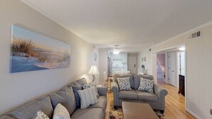 Sala de estar com dois sofás, uma mesa de centro e um quadro na parede. Cozinha visível ao fundo.