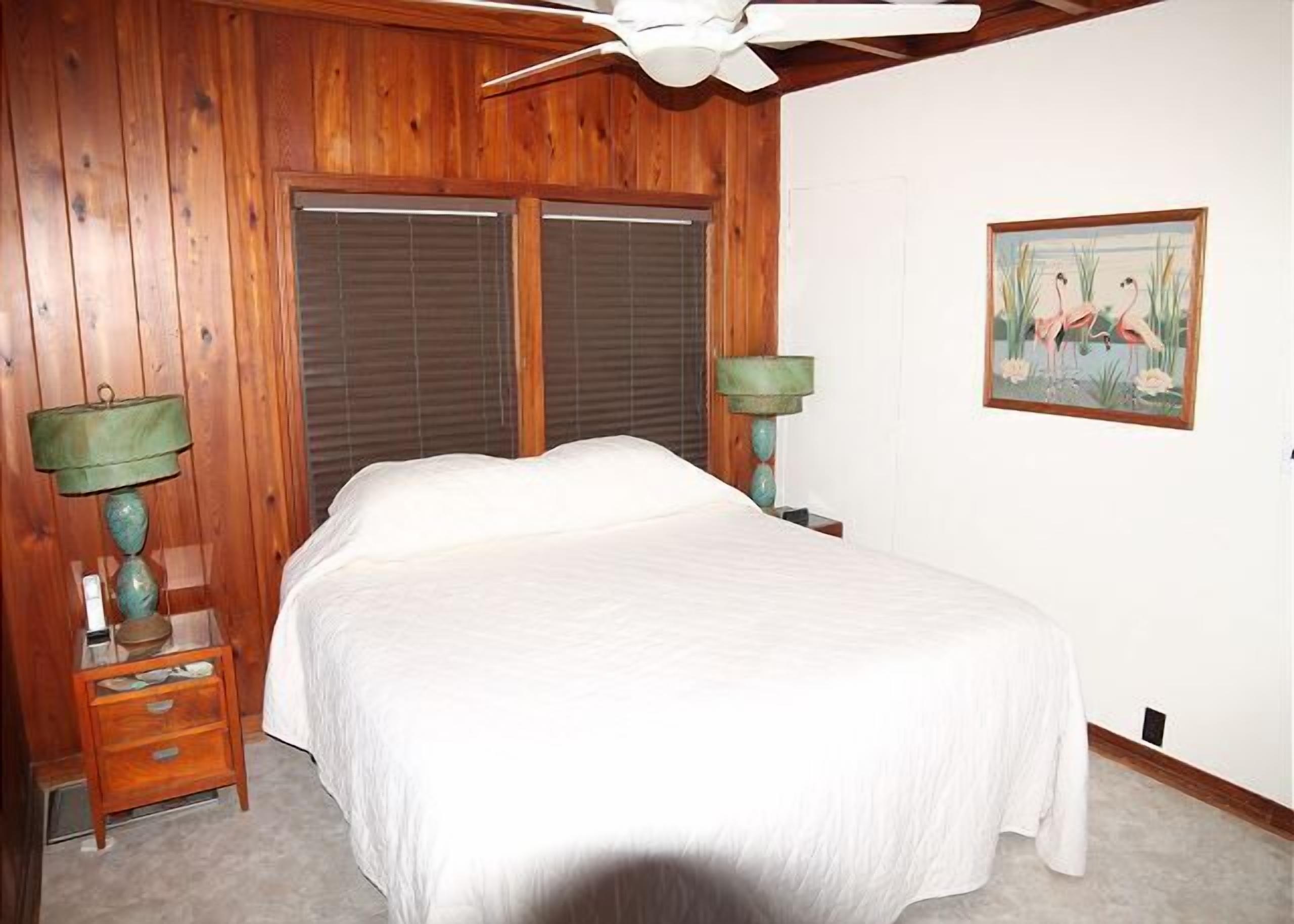 A bedroom with a bed, wooden walls, a ceiling fan, a window with blinds, a nightstand with a lamp, and a painting on the wall.