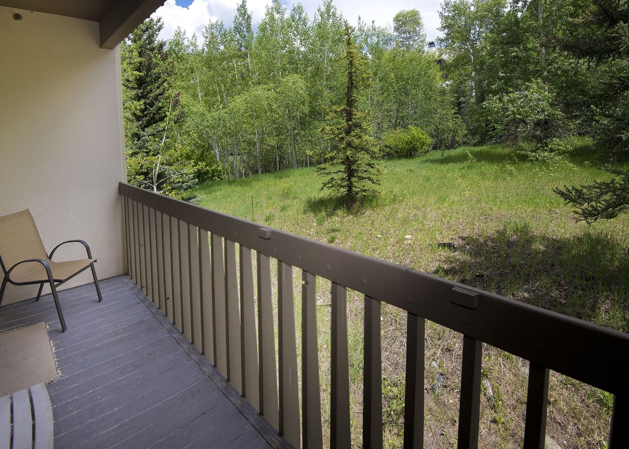 Un balcon doté d’un plancher en bois et d’une rambarde en métal, qui donne sur un espace verdoyant avec des arbres et un petit bâtiment visible au loin.