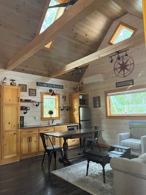 Een knus keuken met houten kastjes, een eettafel en een koelkast. Twee ramen zorgen voor natuurlijk licht en er hangt een plafondventilator.