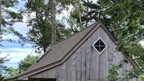 A rustic wooden cabin with a porch, a single window, and an air conditioning unit. - Next Avail. Aug 22 - 3 night min - AC - Premier Cottage, King Bedroom, Pets Free (Round Pond)