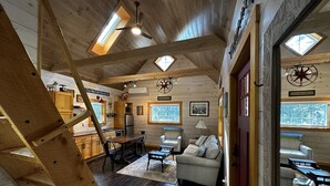 A spacious living area with a high ceiling, wooden beams, and a cozy fireplace. There is a dining area with a table and chairs, and a kitchen in the background. - Next Avail. Aug 22 - 3 night min - AC - Premier Cottage, King Bedroom, Pets Free (Round Pond)